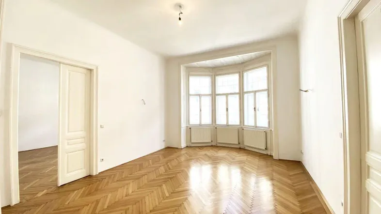 Helles Zimmer mit Fischgrätparkett und einem schönen Erkerfenster, das viel Tageslicht hereinlässt.