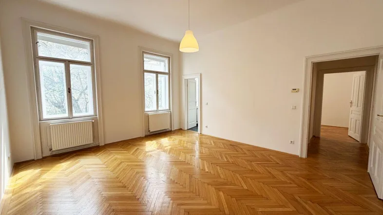 Helles Wohnzimmer mit Fischgrätparkettboden und hohen Fenstern, das viel Tageslicht hereinlässt.