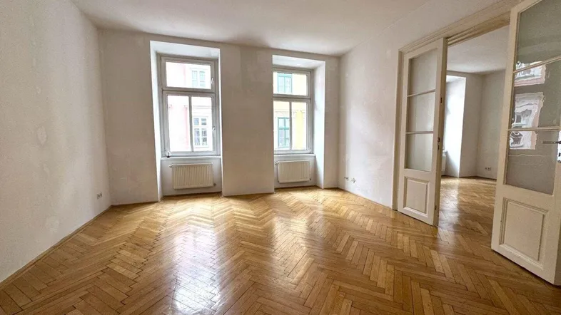 Geräumiges Wohnzimmer mit Fischgrätparkett, hohen Decken und großen Fenstern, die viel Licht hereinlassen.
