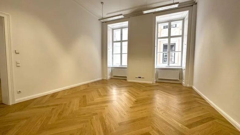 Geräumiger Büroraum mit hellem Fischgrätparkett, hohen Decken und großen Fenstern, die viel Tageslicht hereinlassen.