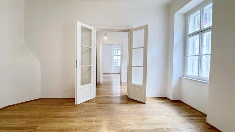 Helles Zimmer mit Holzboden und hohen Fenstern, das durch Flügeltüren mit weiteren Räumen verbunden ist.