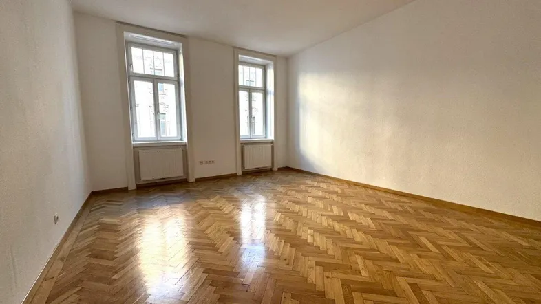 Helles Zimmer mit Fischgrätparkett und zwei Fenstern, ideal als Wohn- oder Schlafzimmer nutzbar.