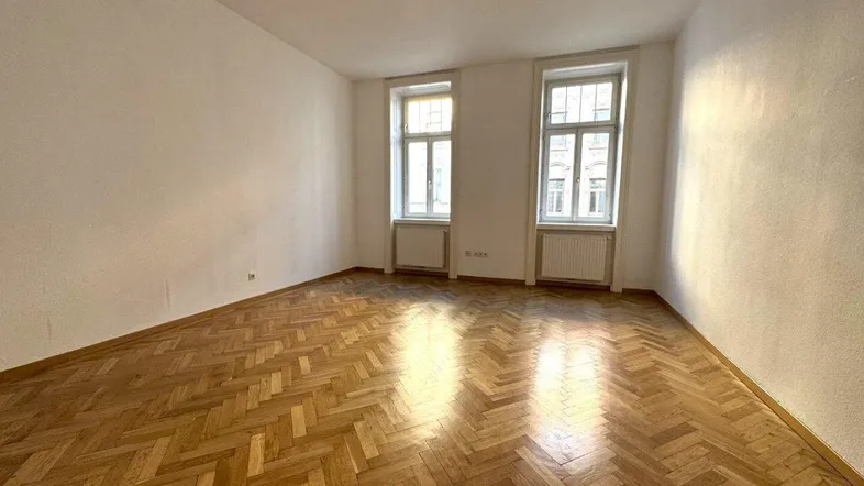 Helles Zimmer mit Fischgrätparkett und zwei Fenstern, bereit zur individuellen Gestaltung.