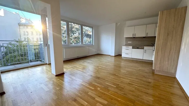 Helles Wohnzimmer mit Holzboden, großen Fenstern, integrierter Küchenzeile und Zugang zum Balkon.