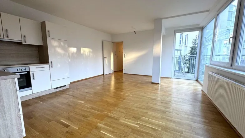 Heller offener Wohnbereich mit integrierter Küche, Parkettboden und Zugang zum Balkon.