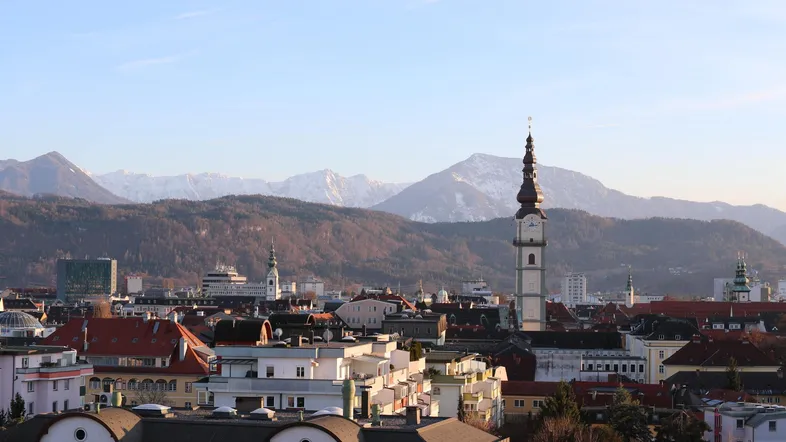 Weitläufige Stadtansicht mit schneebedeckten Bergen im Hintergrund und einem markanten Kirchturm.