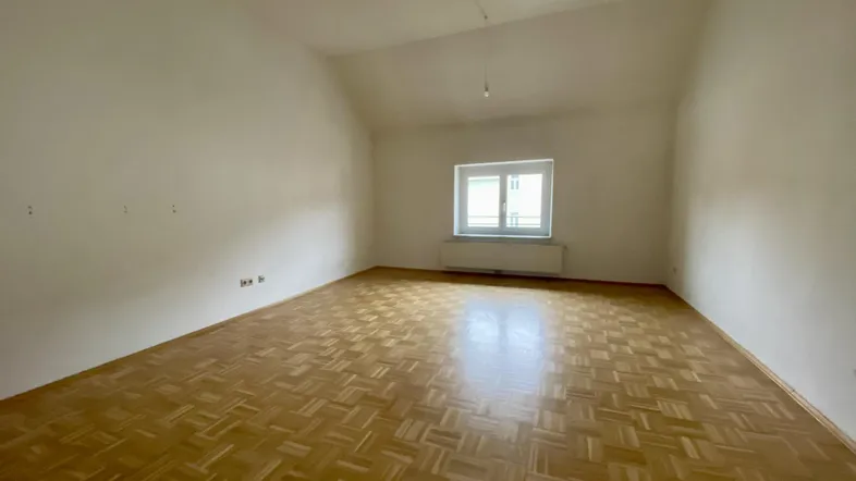 Helles, unmöbliertes Zimmer mit Parkettboden und einem Fenster, das viel Tageslicht hereinlässt.
