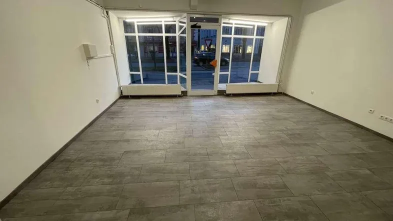 Geräumiger, leerer Raum im Erdgeschoss mit großen Fenstern zur Straße und modernem Fliesenboden.