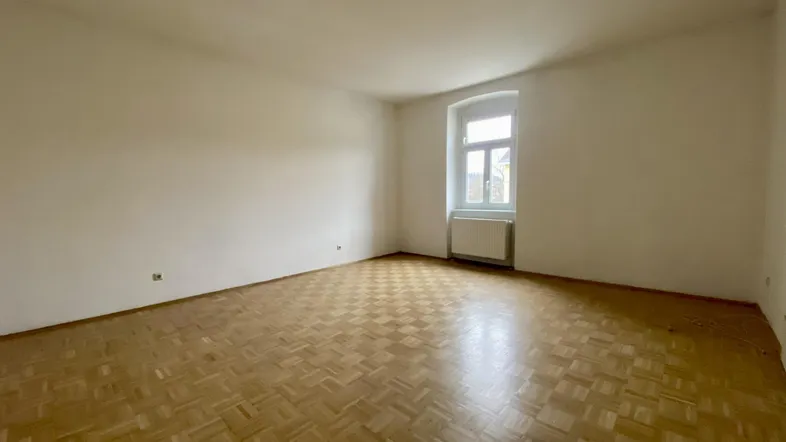 Heller, leerer Raum mit Fischgrätparkettboden und einem Fenster, das Tageslicht hereinlässt.