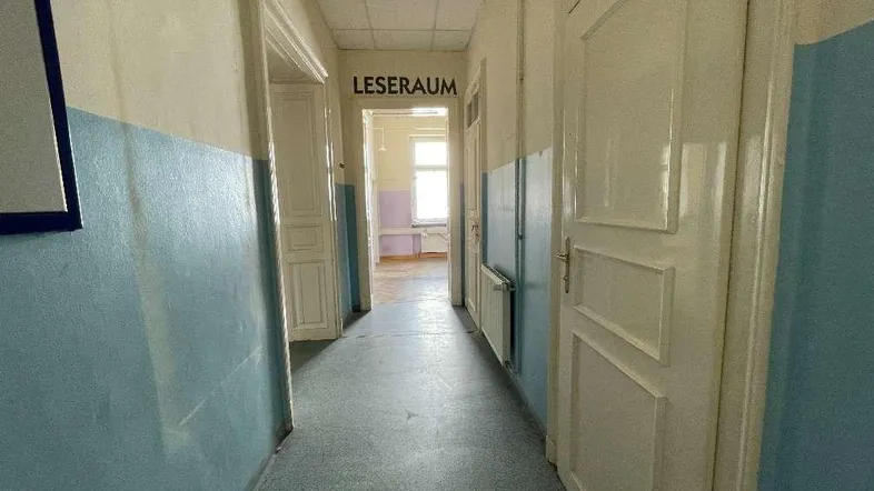 Langer Flur mit zweifarbigen Wänden und Türen, der zu einem Leseraum führt und Renovierungsbedarf zeigt.