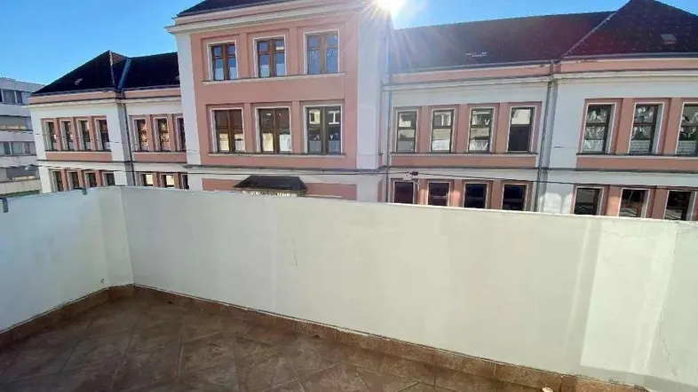 Sonnige Terrasse mit Fliesenboden und Blick auf ein gegenüberliegendes Gebäude mit vielen Fenstern.