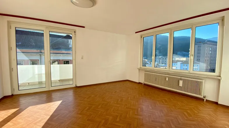 Helles Wohnzimmer mit Fischgrätparkett, großzügigen Fenstern und Zugang zum Balkon mit Fernblick.