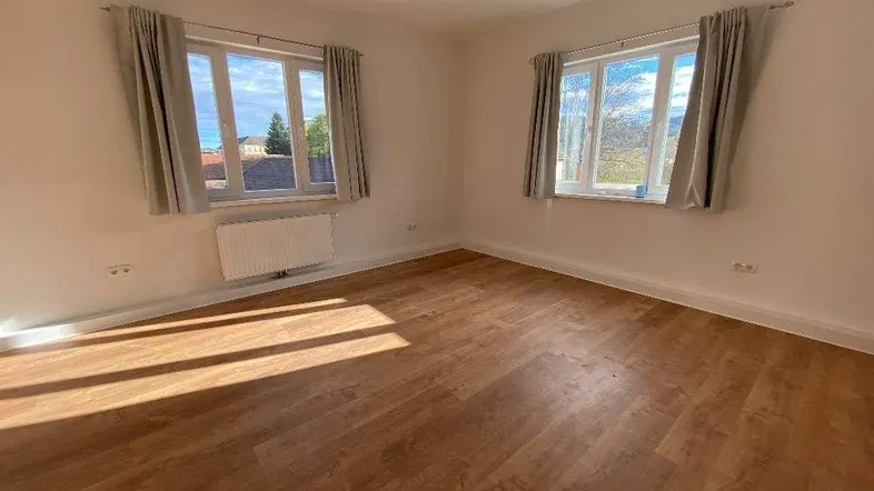 Heller Raum mit Laminatboden und zwei großen Fenstern, die viel Tageslicht hereinlassen.