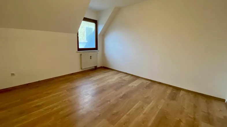 Helles Zimmer im Dachgeschoss mit Holzboden und einem Fenster, das viel Tageslicht hereinlässt.