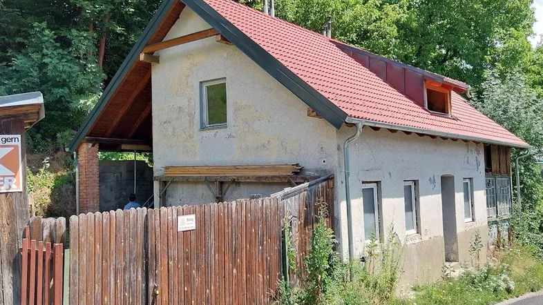 Einfaches Wohnhaus mit rotem Ziegeldach und Holzzaun, umgeben von viel Grün.