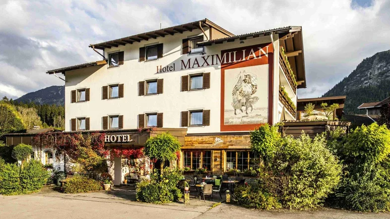 Traditionelles Hotelgebäude mit alpiner Architektur, umgeben von grüner Landschaft und Bergen im Hintergrund.