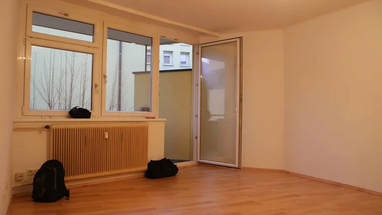Helles Zimmer mit Parkettboden, Heizkörper und Zugang zu einem kleinen Balkon.