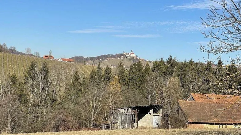 Landschaftliche Ansicht mit Hügeln, Wäldern und einem Dorf in der Ferne unter blauem Himmel.