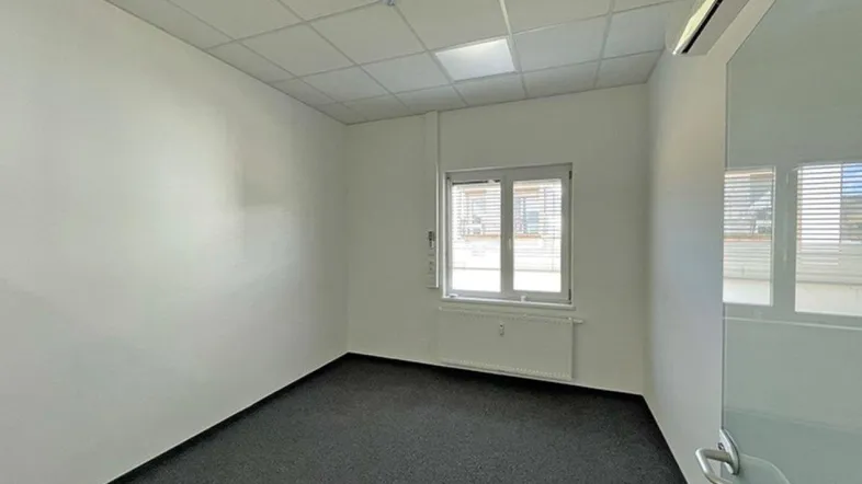 Leeres Büro mit weißen Wänden, dunklem Teppichboden, Fenster und Klimaanlage an der Decke.