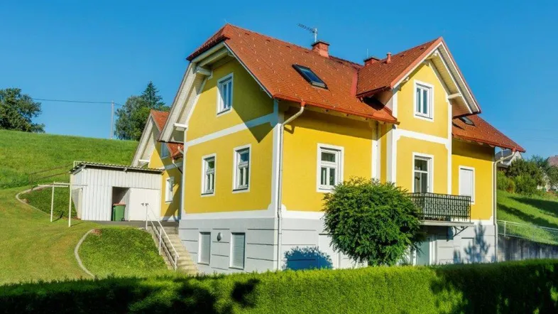 Gelbes Einfamilienhaus mit rotem Satteldach, kleinem Balkon und gepflegtem Gartenbereich in ländlicher Umgebung.