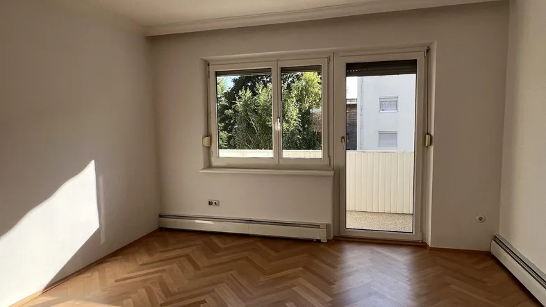 Heller Raum mit Fischgrätparkettboden und Zugang zu einem sonnigen Balkon.