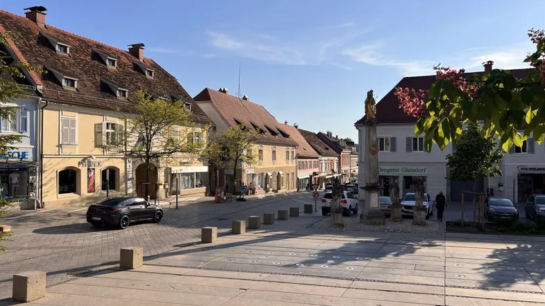 Blick auf einen belebten Stadtplatz mit historischen Gebäuden und geparkten Autos unter blauem Himmel.
