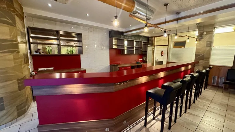 Eine moderne Bar mit roter Theke, Holzverkleidung und Barhockern, beleuchtet von Hängelampen.