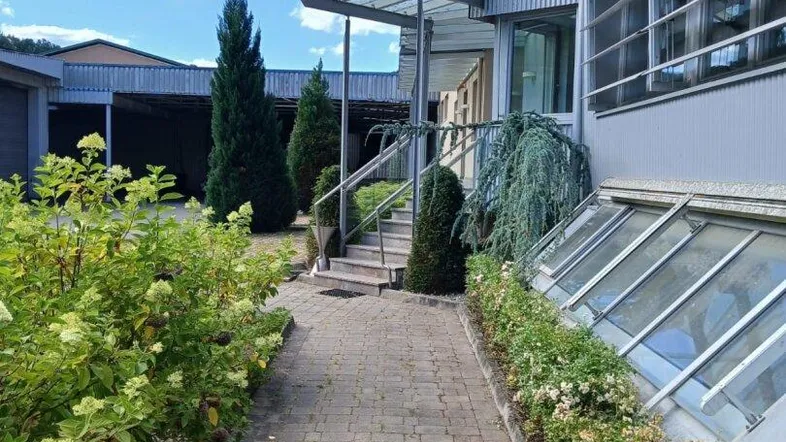 Eingangsbereich eines Gebäudes mit gepflastertem Weg, Treppe und Vordach, umgeben von Grünflächen unter blauem Himmel.