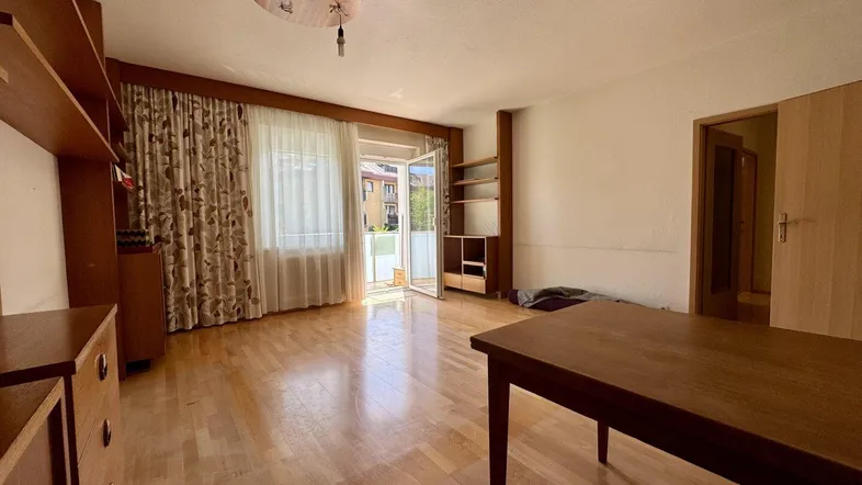 Helles Wohnzimmer mit Parkettboden, Holzmöbeln und Zugang zu einem Balkon mit Blick auf die Umgebung.