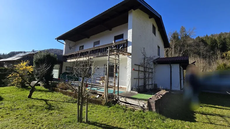 Weißes Haus mit Balkon und Terrasse, umgeben von Grünflächen und Bäumen unter blauem Himmel.