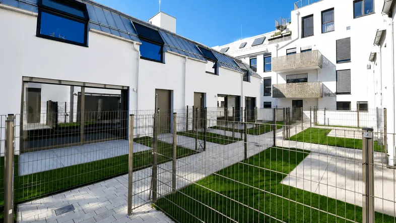 Moderne Wohnanlage mit weißen Fassaden, privaten Gärten und gepflasterten Wegen unter blauem Himmel.