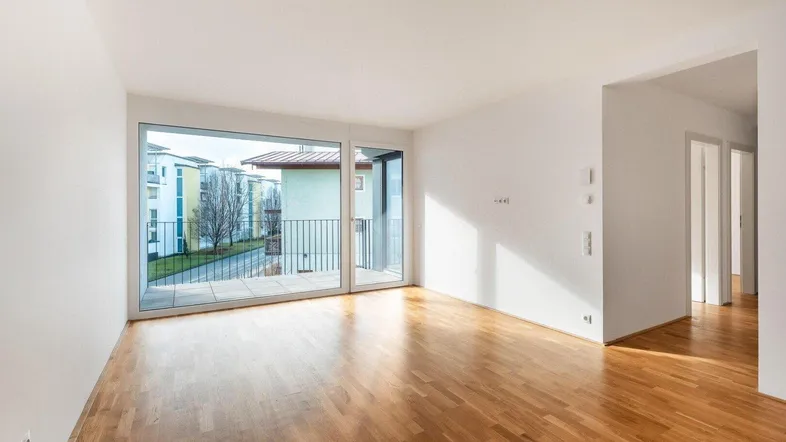 Helles Wohnzimmer mit Parkettboden und großem Fenster, das Zugang zu einem Balkon bietet.