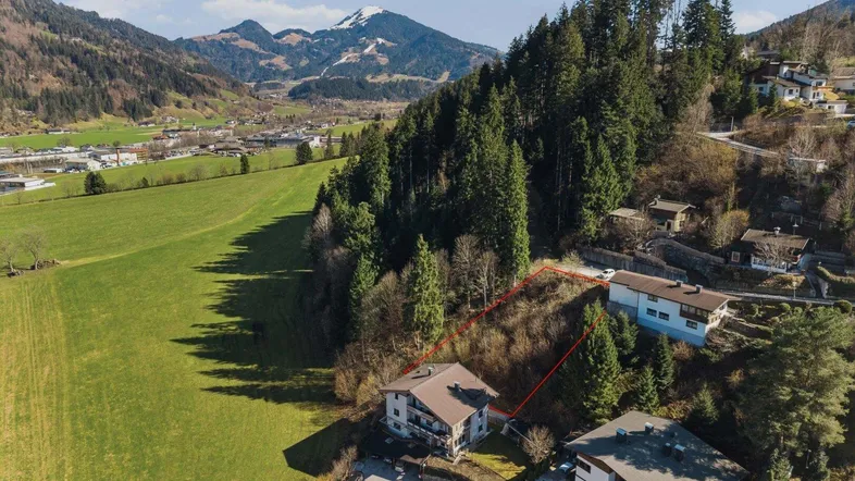 Luftaufnahme eines Baugrundstücks am Hang mit umliegenden Häusern und Bergblick.