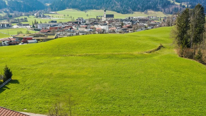 Weitläufige grüne Wiesen mit einem malerischen Dorf und Bergen im Hintergrund, ideal für Naturliebhaber.