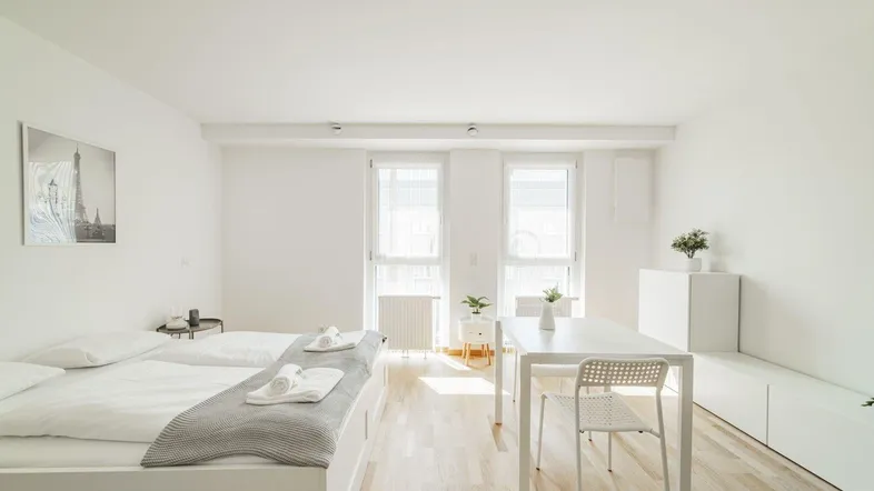 Helles Studio-Apartment mit Doppelbett, Esstisch und großen Fenstern, modern eingerichtet.