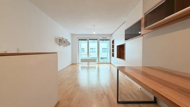 Heller Wohnbereich mit Parkettboden, integrierten Regalen und Zugang zum Balkon.