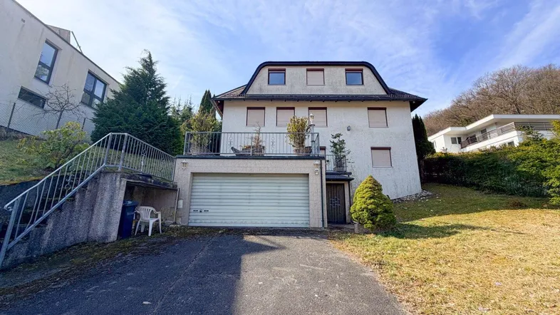 Weißes Einfamilienhaus mit Garage und kleinem Balkon, umgeben von grüner Landschaft.