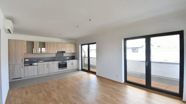 Heller offener Wohnbereich mit integrierter Küche, Parkettboden und Zugang zum Balkon.