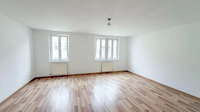 Helles, leeres Zimmer mit Laminatboden und zwei Fenstern, bereit zur individuellen Gestaltung.