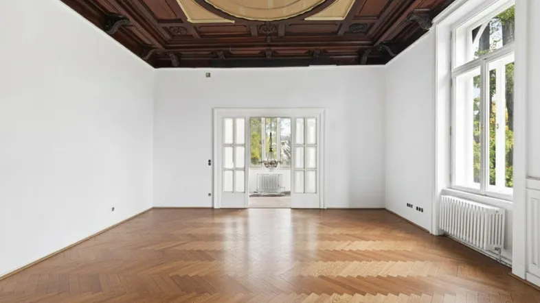 Geräumiges Zimmer mit beeindruckender Holzkassettendecke, Fischgrätparkett und großen Fenstern.