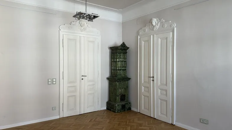 Geräumiges Zimmer mit Fischgrätparkett, hohen Decken und einem traditionellen grünen Kachelofen.
