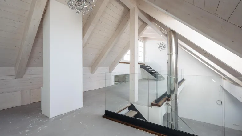 Heller Dachbodenraum mit sichtbaren Holzbalken, moderner Beleuchtung und einer eleganten Glasbrüstung.