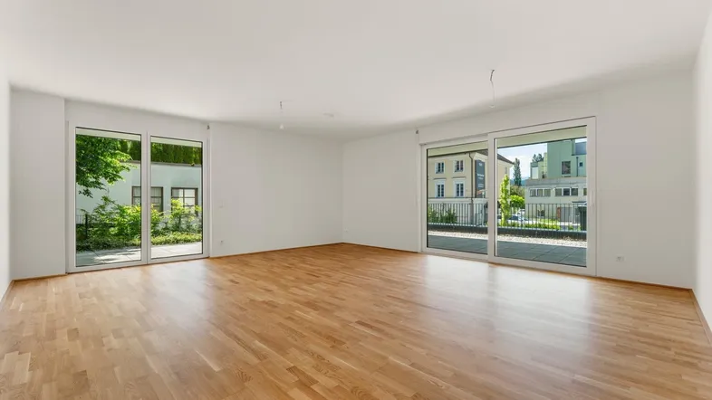 Heller, leerer Wohnraum mit Parkettboden und großen Fenstern, die viel Tageslicht hereinlassen.