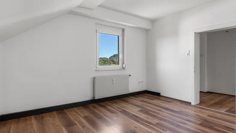 Helles Schlafzimmer im Dachgeschoss mit Laminatboden, schräger Decke und Ausblick ins Grüne.