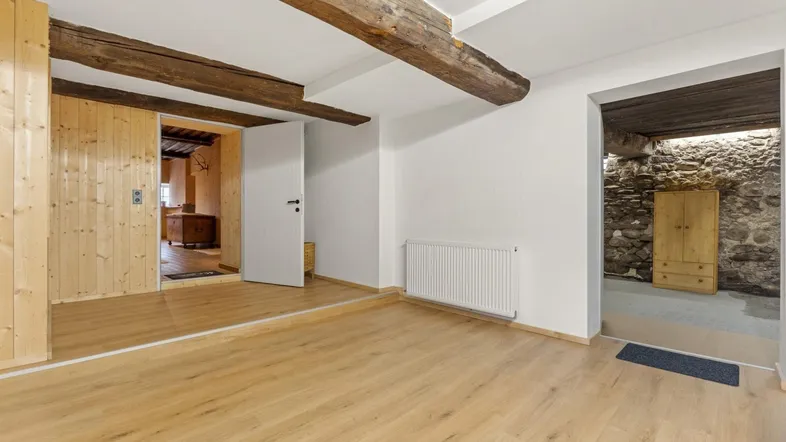 Geräumiger Wohnbereich mit Holzbalken, Parkettboden und Zugang zu einem rustikalen Raum.