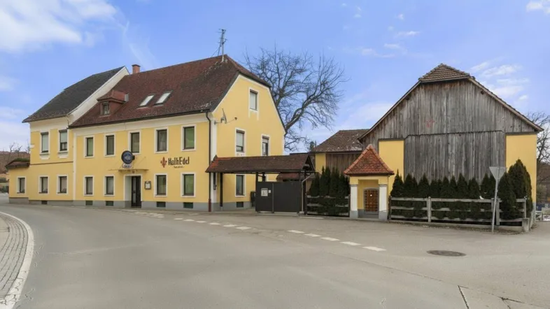 Ein traditionelles gelbes Gebäude mit rotem Ziegeldach und einem angrenzenden Holzschuppen, gelegen an einer Landstraße.