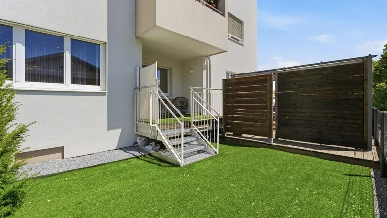 Ebenerdige Terrasse mit Kunstrasen und Zugang zum Gebäude, umgeben von einer Holzwand.