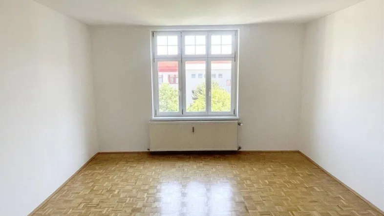 Helles, leeres Zimmer mit Parkettboden und einem großen Fenster, das viel Tageslicht hereinlässt.
