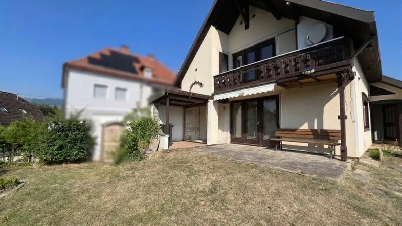 Traditionelles Haus mit Balkon, gepflasterter Terrasse und großem Gartenbereich unter blauem Himmel.