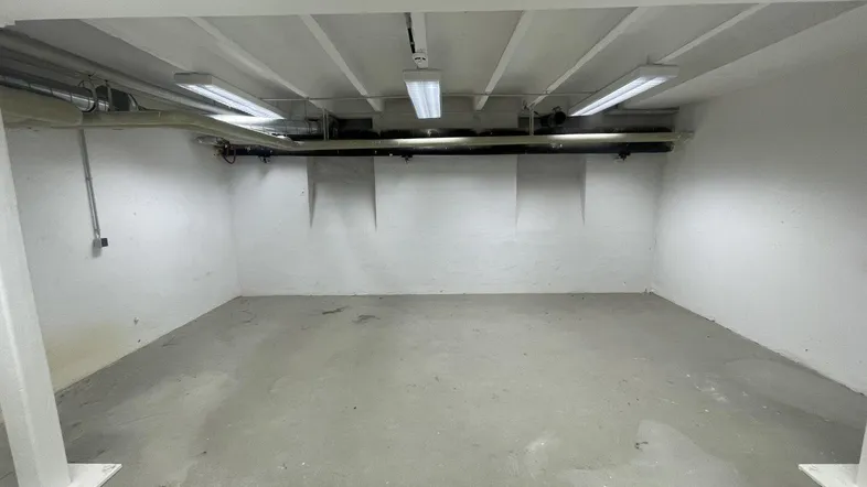 Leerer Kellerraum mit Betonboden, weißen Wänden und sichtbaren Installationen an der Decke.